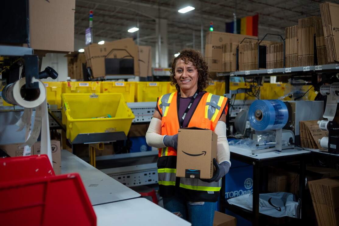 Trabajadora de Amazon cargando una caja delante en una estación de empaque. 