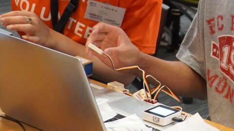 机上に開かれたノートパソコンのキーボード部分に、白いデバイスや紙片が置かれている。グレーのTシャツの人物がデバイスにつながった白い部品を手に持ち、オレンジ色のTシャツの人物が隣に座っている。