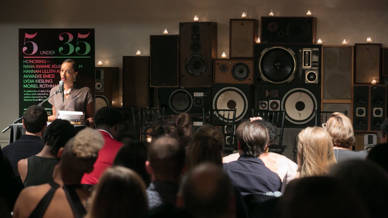 一位女士站在讲台上,对着麦克风讲话。她面前坐着一群人,面对着她。