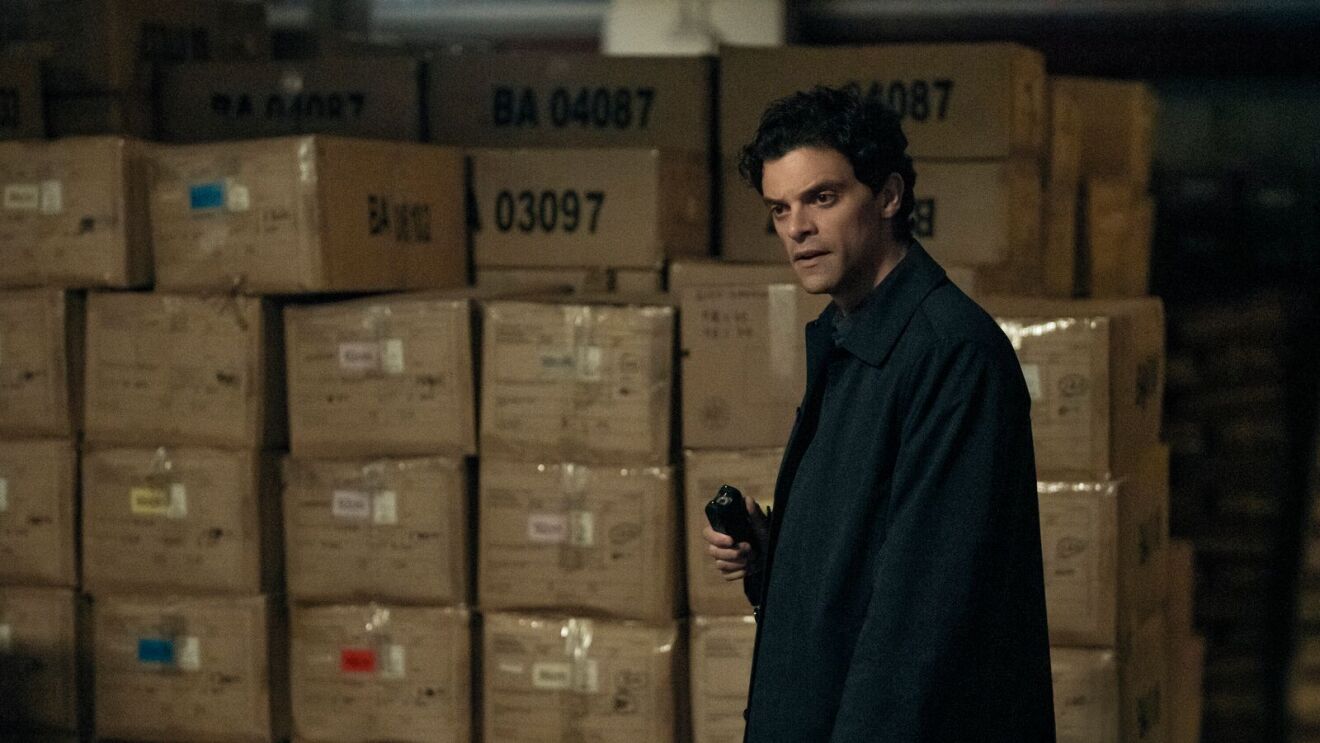 DCI Rhys (Jacob Fortune-Lloyd) in dark coat standing amid stacks of numbered shipping boxes