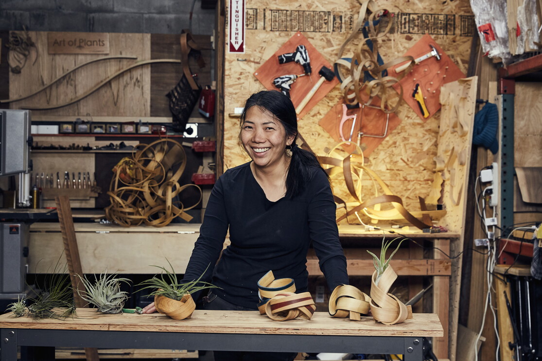 A woman stands behind a work table, smiling at the camera. In front of her are several handmade bentwood air plant containers, behind her sits more wood and tools. 