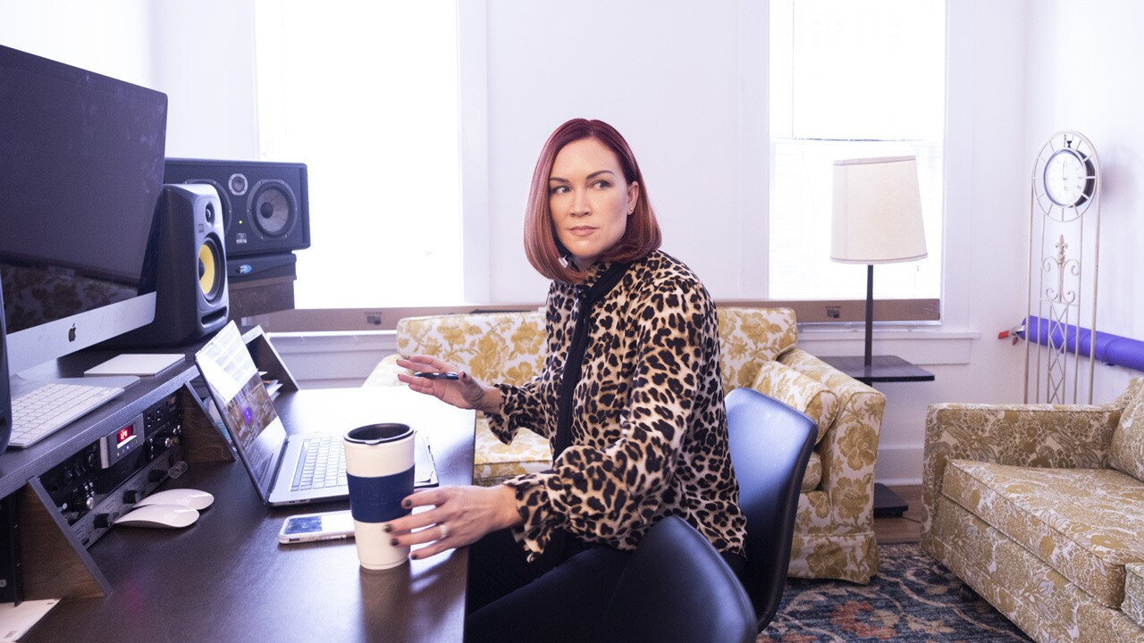 An image of MTK working at a desk with large speakers surrounding her. She is looking behind her and reaching for a coffee next to her on the desk.