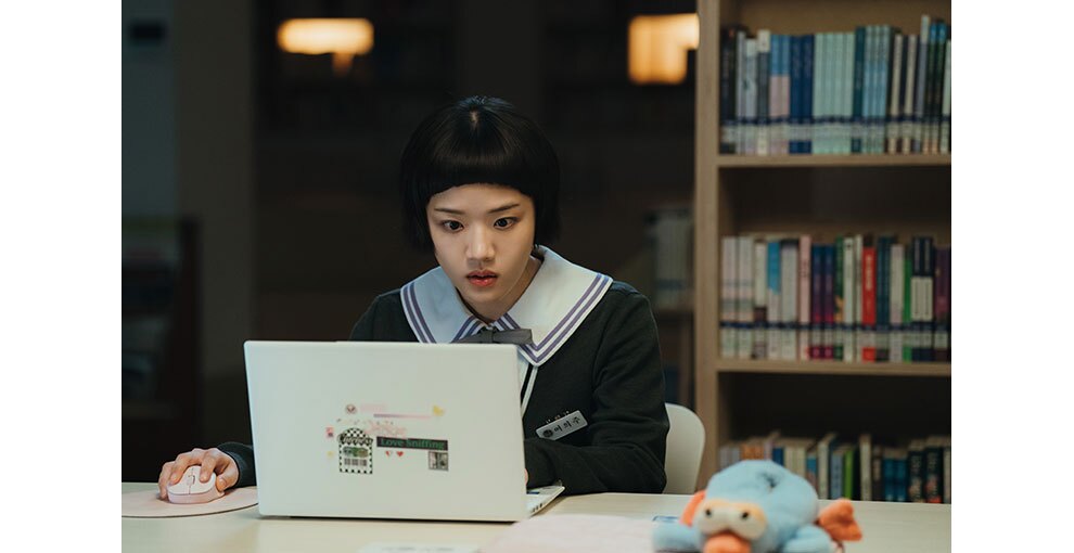 図書室で机に向かい、ノートパソコンの画面を見つめる少女。襟の大きな制服を着用。机の右側にぬいぐるみ。