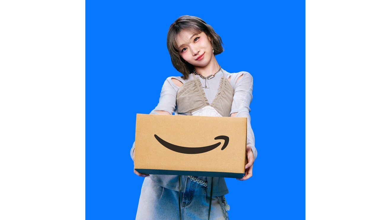 若い女性が、両手でAmazonロゴ入りダンボール箱を持って前に差し出している。