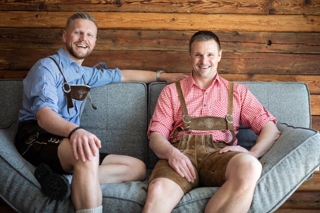 Zwei Männer in Lederhosen sitzen auf einem grauen Sofa und lachen in die Kamera.