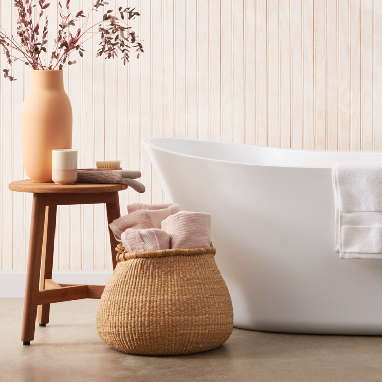 Foto de un baño al estilo bogegón. La pared es imitación madera con tablas verticales de color blanco roto. Una bañera de color blanco de las que no están pegadas a la pared. Con una toalla blanca apoyada. En el centro, un saco de mimbrte con tres toallas rosas  sobresaliendo. Y a la izquierda un taburete con un vaso de color blanco y rosa, una toalla de color marrón, un esponja de baño estilo cepillo y un jarrón de barro  con su color original con unas hojas secas de color morado oscuro. 