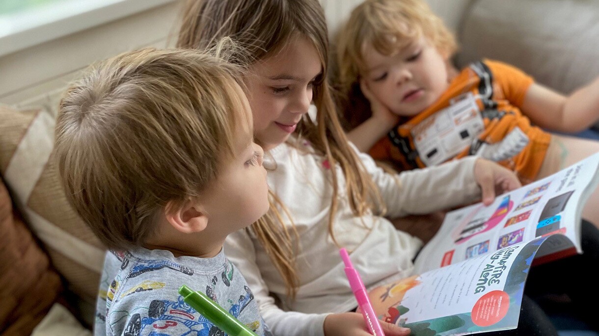 Three children read Amazon's holiday toy catalog together, holding markers to circle the items they want.