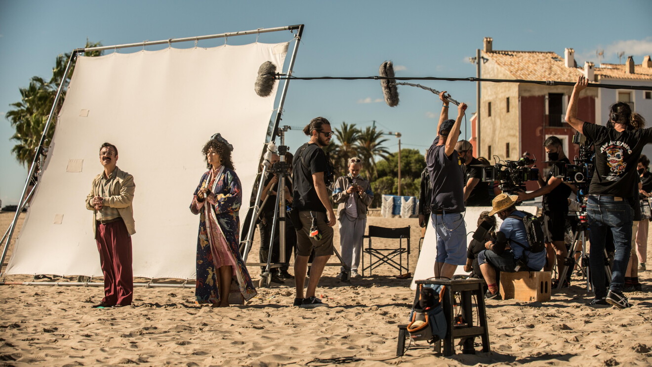 En un set de rodaje exterior, en la arena de la playa, aparecen dos actores, hombre y mujer vestidos de los años 60. Una pantalla blanca para aportiguar la luz. De fondo dos casa de pueblo marinero y, al lado de los actores, un equipo técnico de unas 10 personas con cámaras y micros. Todos van vestidos con ropa oscura. 