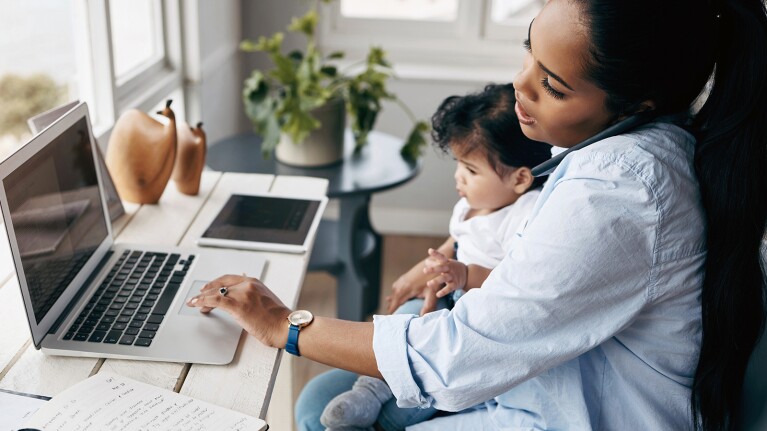 Image of a woman on the phone and working on a computer while holding a baby.