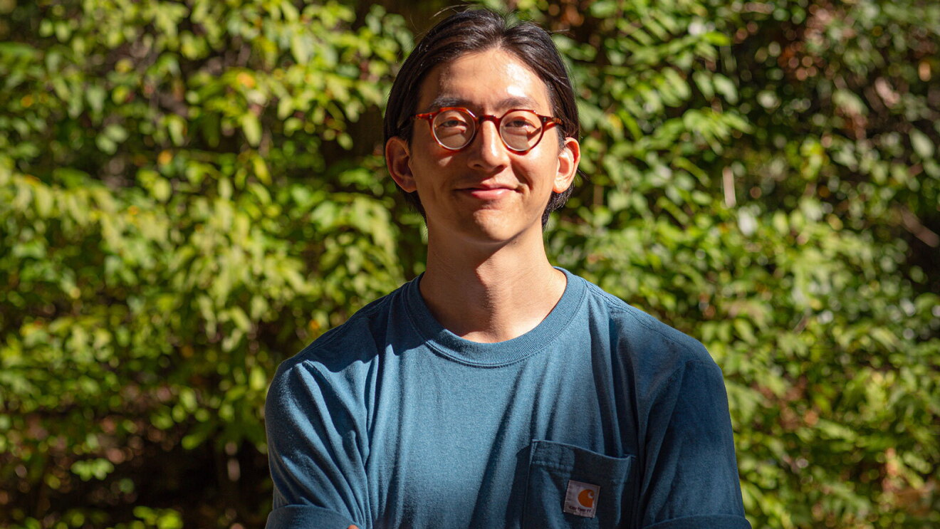 An image of a man smiling for a photo outside in front of a large green shrub.