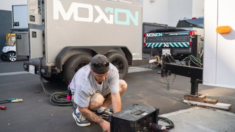 Man in casual clothes bending down to adjust some technical equipment in the backlot of a movie studio. Behind him is a Moxion truck and trailer.