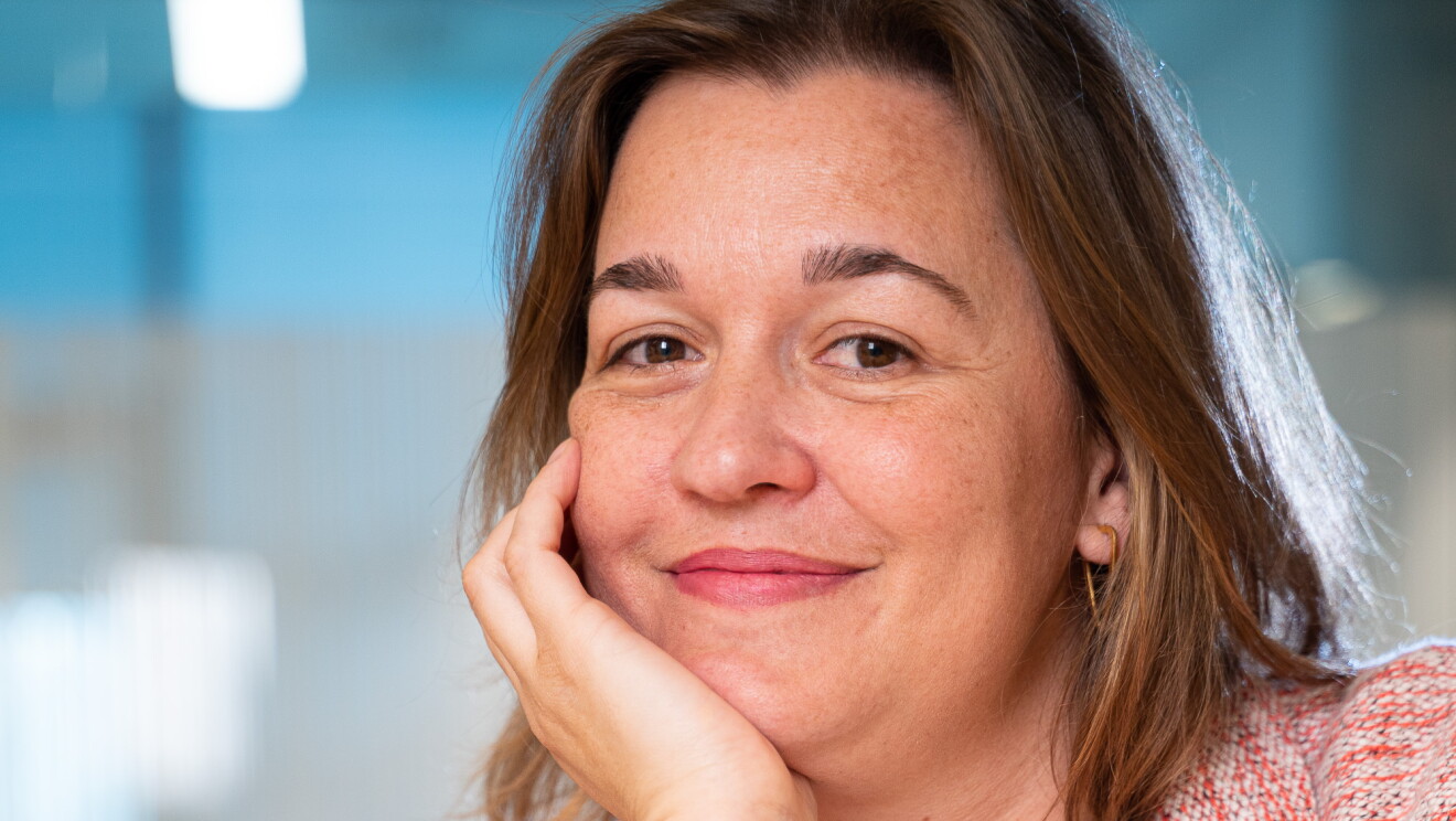 Marta Fernández, Content Program Manager de Alexa delante de un dispositivo Amazon Echo. Está apoyada en una mesa con un brazo en la mejilla. LLeva una camiseta blanca y una chaqueta rosa. Además lleva un collar de color de oro. Tiene media melena rubia. 