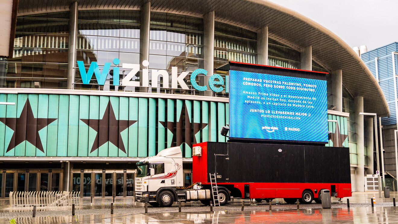 #SeriesDesdeMiBalcón es una iniciativa creada por Amazon Prime Video, el ayuntamiento de Madrid y Exterior Plus. Es la plaza Felipe II de Madrid. De fondo el palacio de deportes Wizink Center. Justo delante un trailer de colo blanco con una caja negra y roja. Encima una pantalla gigante donde se proyecta la serie.