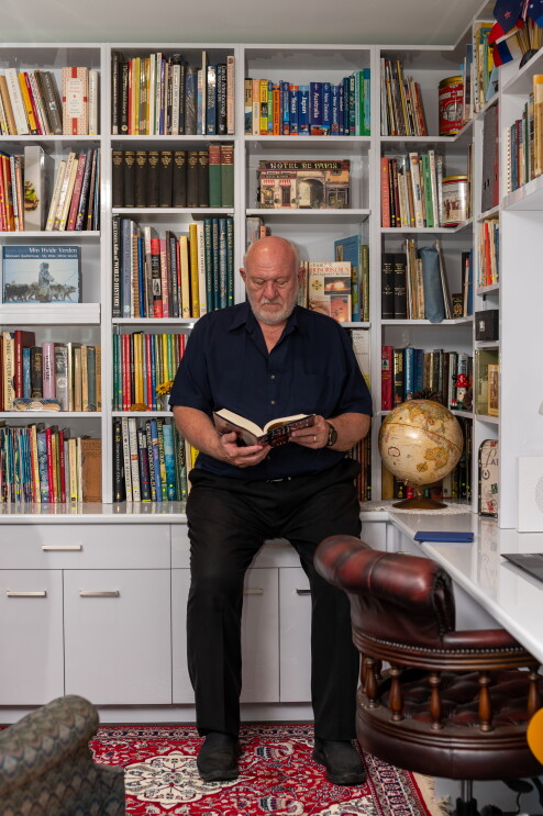 An older, distinguished man sits on his book shelf ledge, while paging through a book he wrote