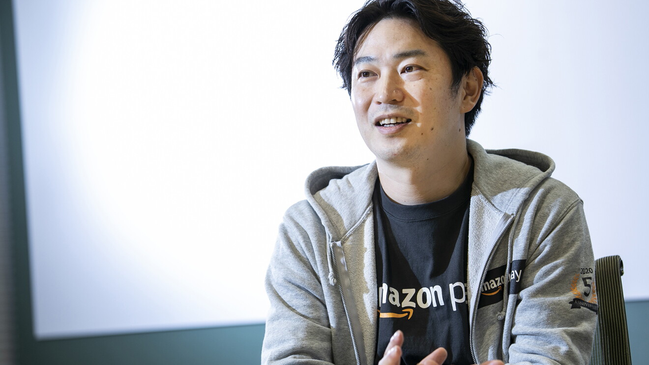 白い壁を背に椅子に座って話す男性。グレーのパーカー、黒いシャツを着用。