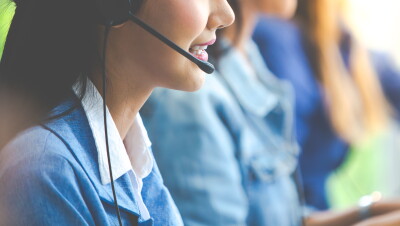 Três mulheres sentadas diante de uma mesa. Em destaque, uma delas está usa um fone com um microfone acoplado