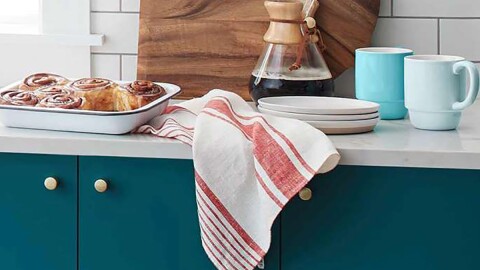 Image from within a kitchen, with cinnamon rolls, a Chemex coffee maker, towel, cutting board, mugs, and plates on the counter. 