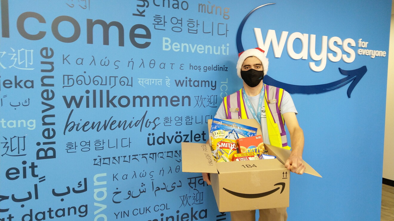 An image of an Amazon employee holding a box of donations. He is standing in front of a wall with the "Wayys" logo on it and is wearing a Santa hat, a face mask, and a safety vest.