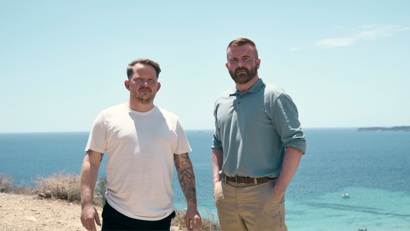 Two stylish, middle-aged men standing infront of ocean, looking into camera