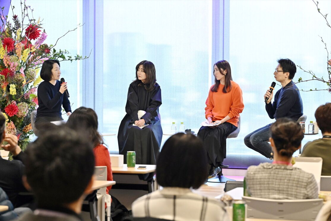 ステージでショートヘアの女性が話していて、2人の女性と1人の男性がその話を聞いている。