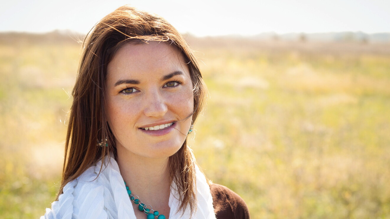 A woman in a brown dress, white scarf, and turquoise necklace in a field in the sun. 