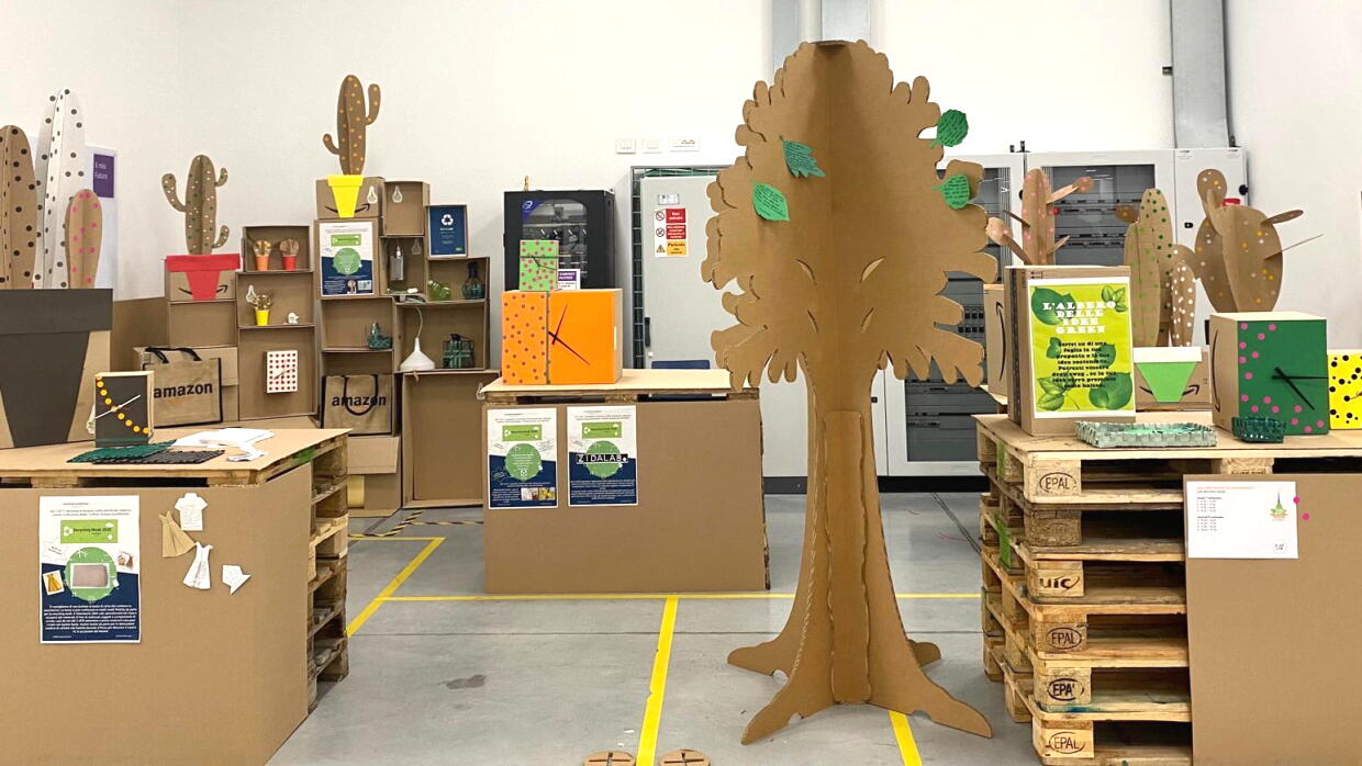 All'interno del centro di distribuzione di Vercelli, area realizzata con materiali riciclati tra cui cartone. Al centro c'è la sagoma di un albero, realizzato in cartone, un corner con piante realizzate in cartone e orologi da parete realizzati in cartone. 