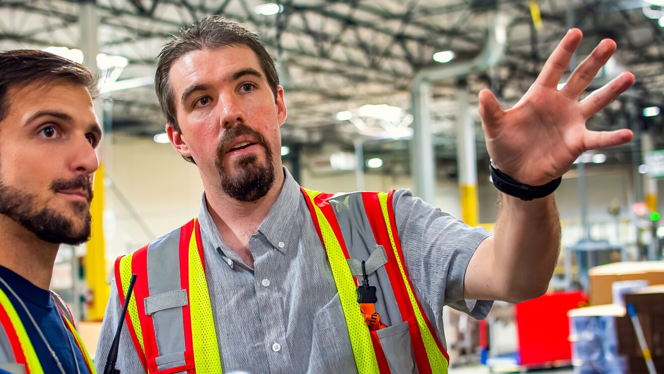 Two men in safety vests. One man gestures while the other looks on.