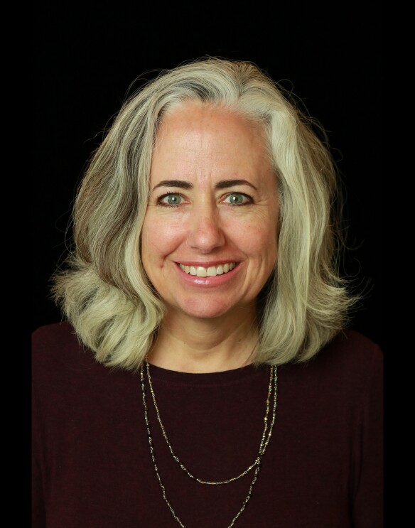 A woman with blonde hair smiles at the camera, she sits in front of a black background.