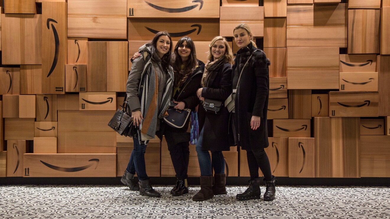 Quattro ragazze sorridono di fronte a un muro di scatole Amazon. Sono Monika, Urte, Francesca e Ankita, vincitrici dell'Amazon Design Award, quattro studentesse del Master in Interior Design dello IED Milano hanno vinto gli Amazon Design Awards, una competizione in cui si chiedeva ai corsisti di ripensare i depositi di smistamento Amazon con uno sguardo alla praticità e al comfort dei dipendenti.