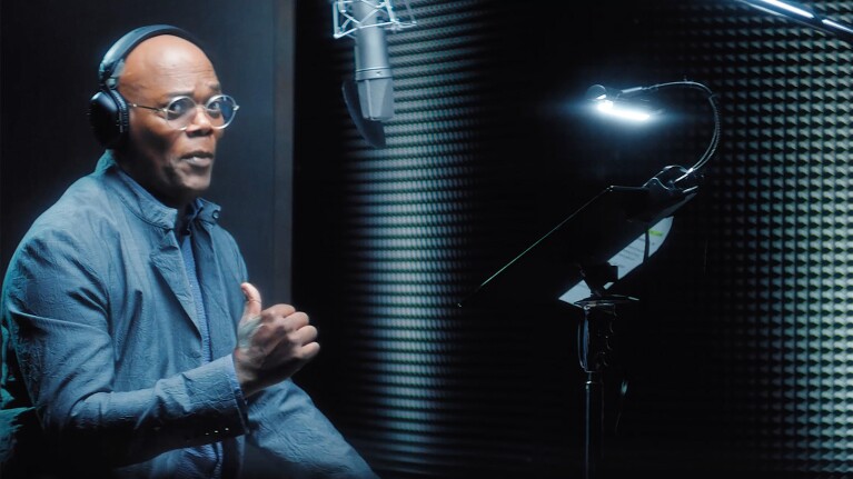 An image of a man recording in a studio wearing a blue/grey jacket and showing a thumbs up. 