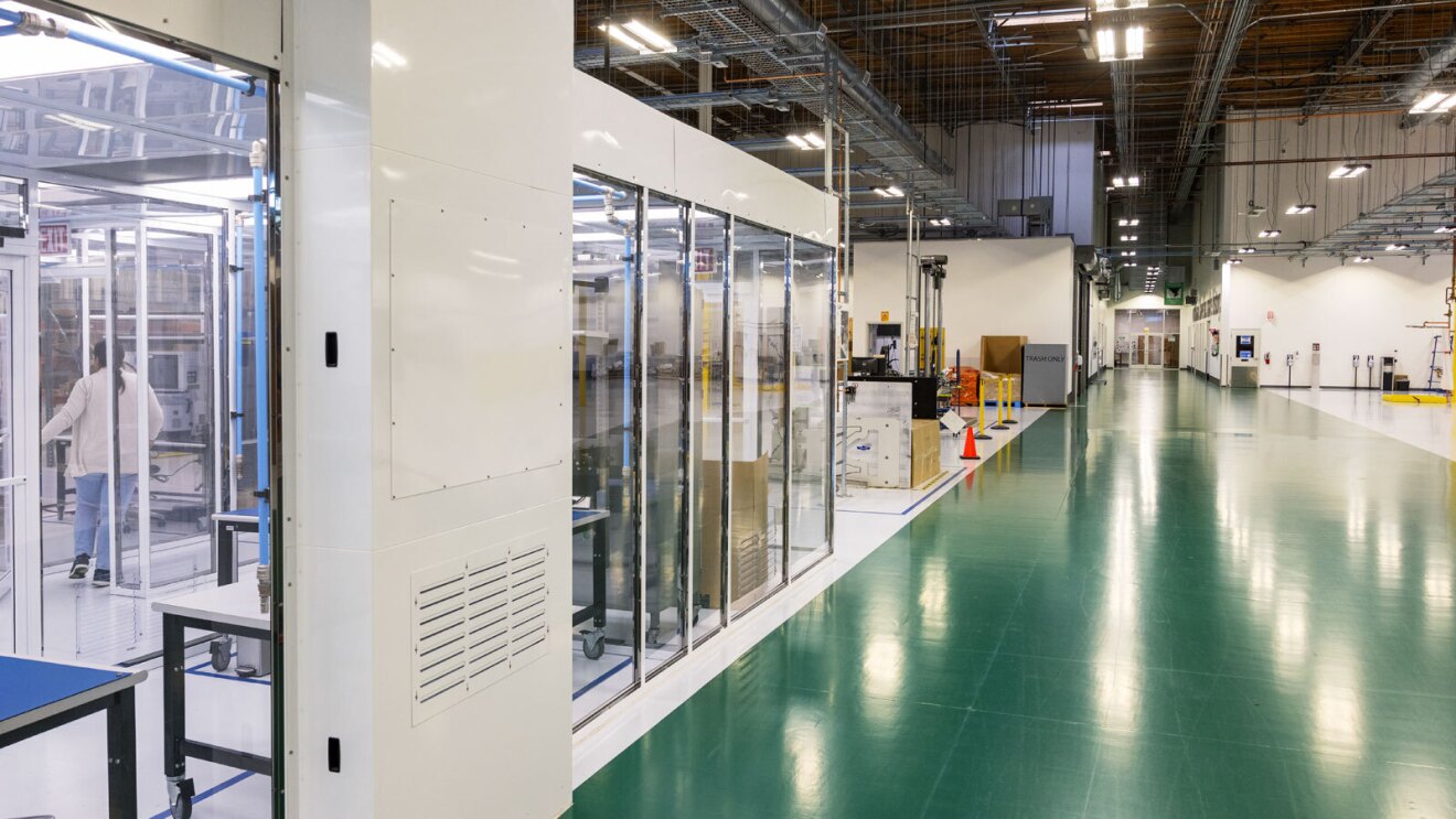 An image of the inside of a large, lab building with a green floor and white walls. There is a glass room area along with open spaces with various machines and boxes lined up.