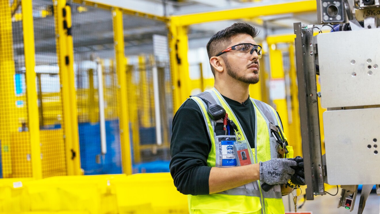 A man wears a yellow safety vest and goggles as he works in a fulfillment center.