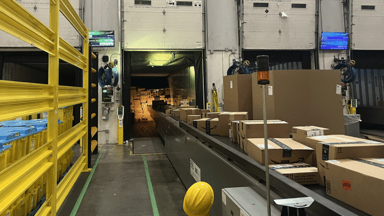 An image of boxes on a conveyer belt in an Amazon fulfillment center leading up to an open truck where there are a lot of boxes stacked.