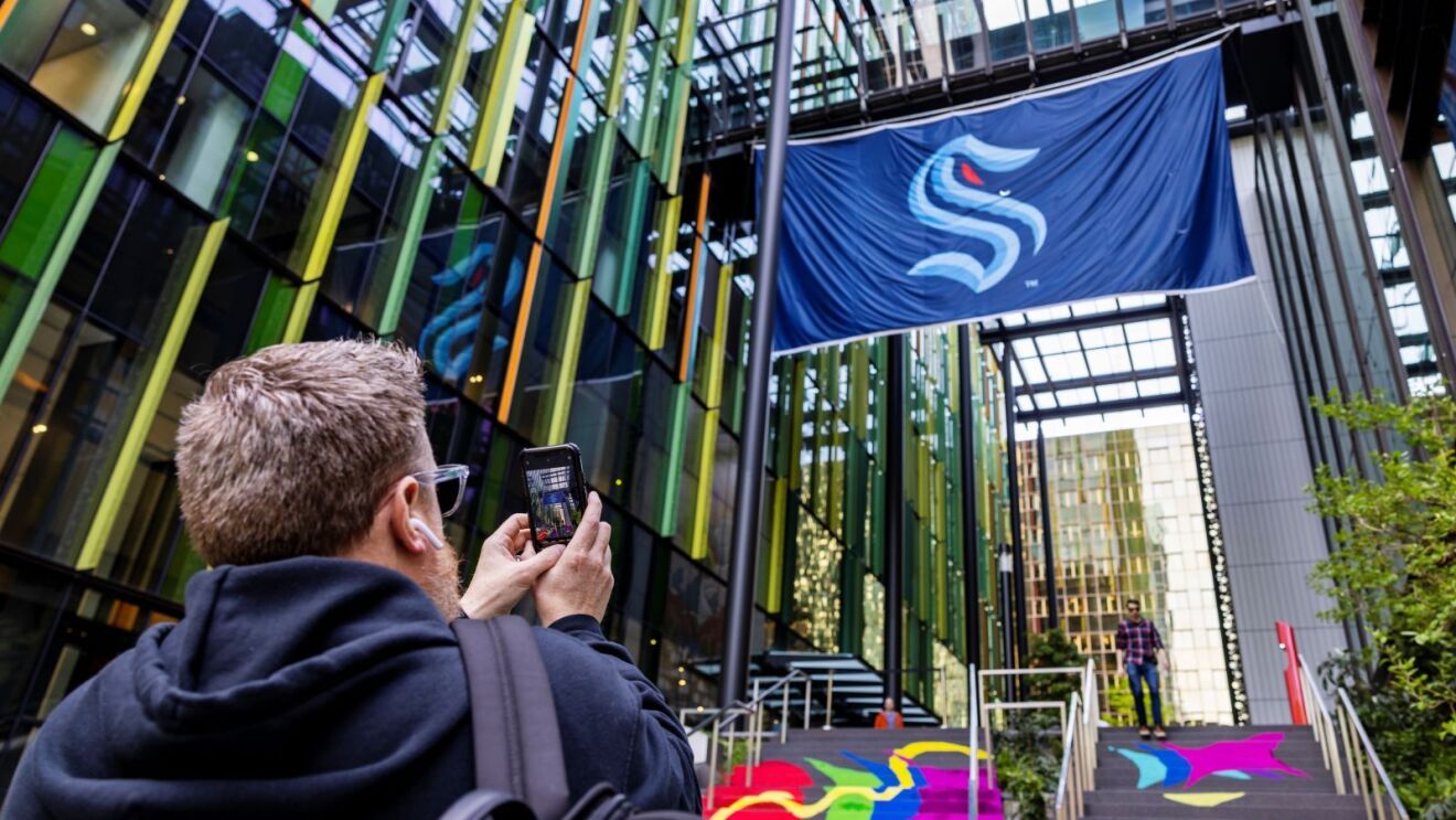 Man holding his phone up, taking a photo of a large Seattle Kraken flag that hangs between two buildings in downtown Seattle.