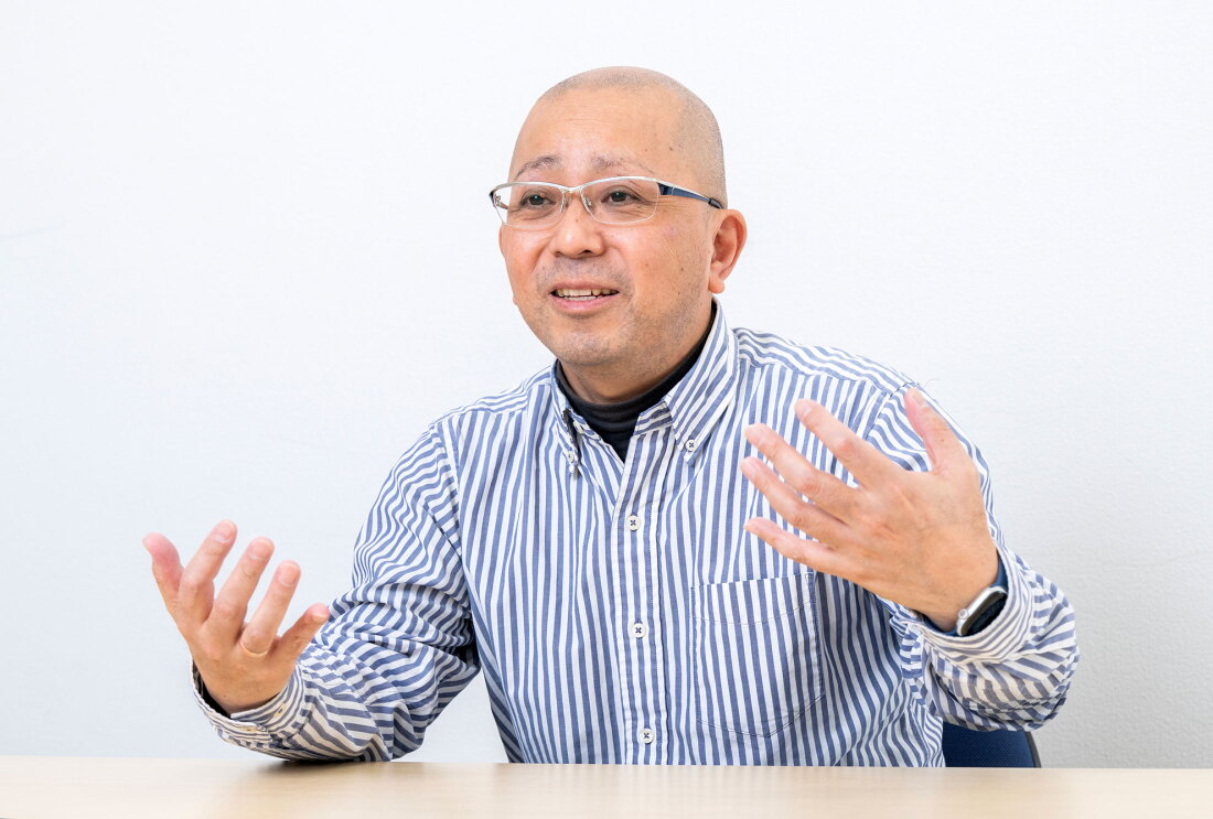 白い壁を背景にテーブルに向かって座る人物。薄い青と白のストライプシャツの下に黒いハイネックを着用し、眼鏡をかけている。両手を体の前で広げてジェスチャーを交えながら話をしている。