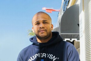 An image of a man standing for a photo outside. There is a palm tree and a street view behind him.