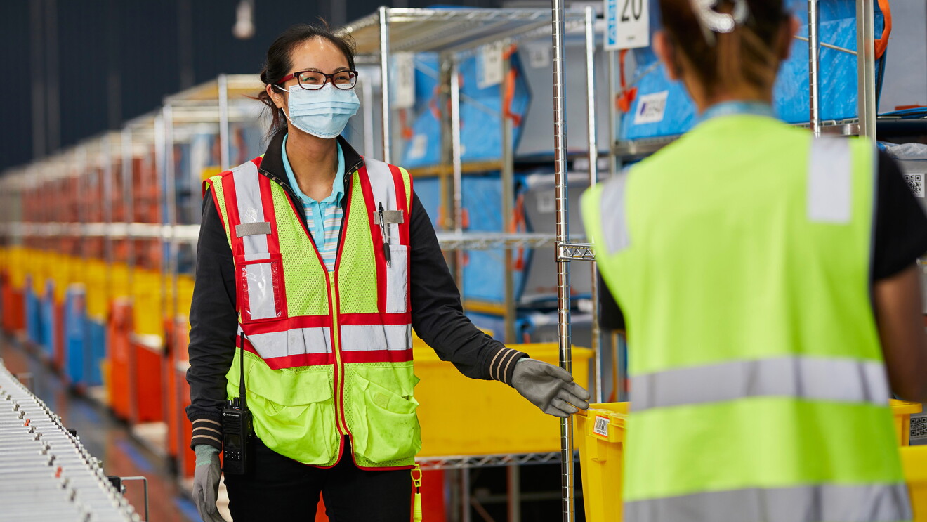 An image of two women working in an Amazon fulfillment centre. They are both wearing safety vests and masks and smiling at each other.
