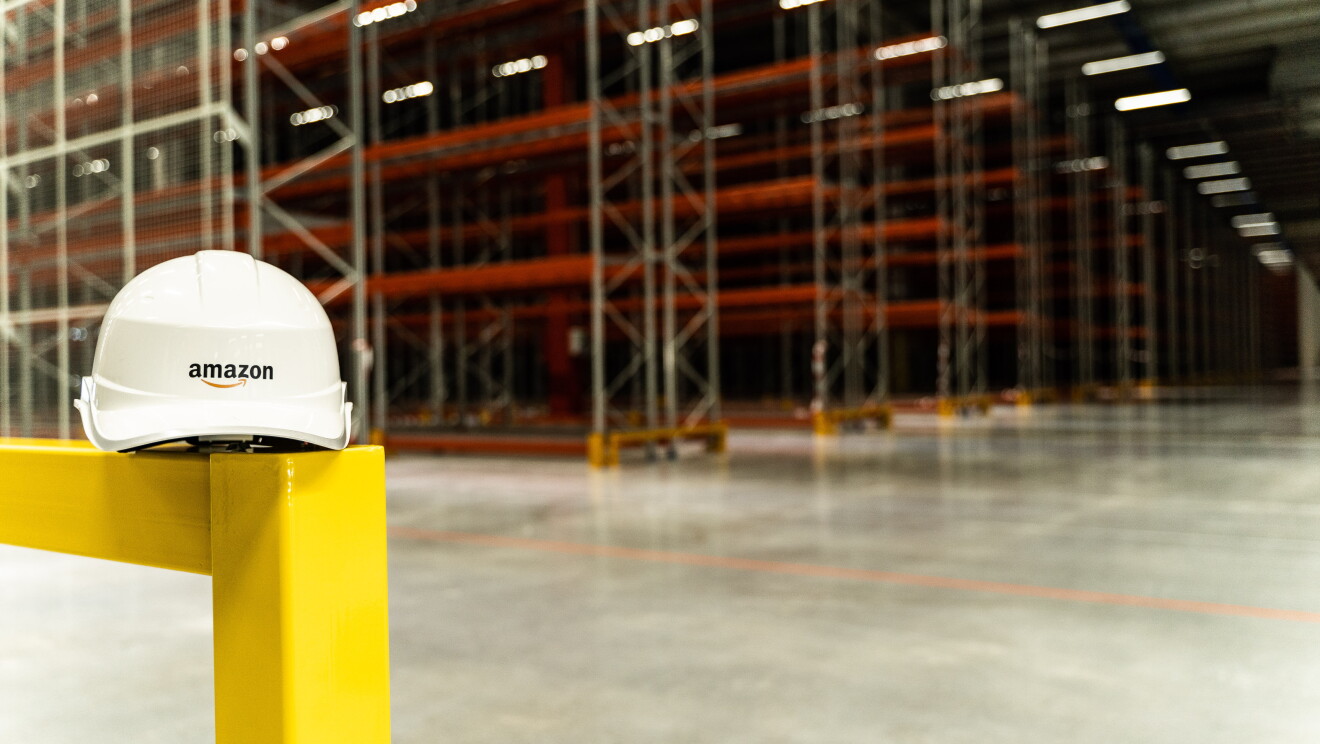 En primer plano aparece un casco de Amazon, de color blanco y con su logo. Está apoyado en una baranda amarilla. De fondo, las estanterías de color rojo. 