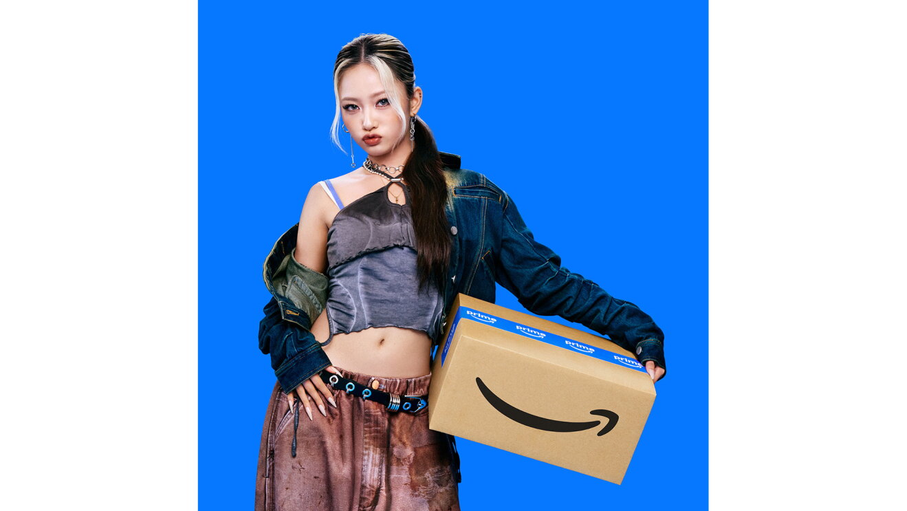 黒髪の一部を白くブリーチした若い女性が、Amazonロゴ入りダンボール箱を左手と腰で挟んで持っている。