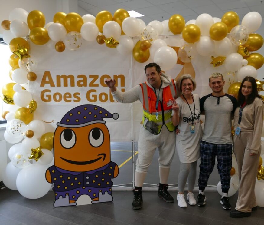 Im Amazon Logistikzentrum Frankenthal stehen vier Mitarbeiter:innen im Pyjama vor dem Banner „Amazon Goes Gold“.