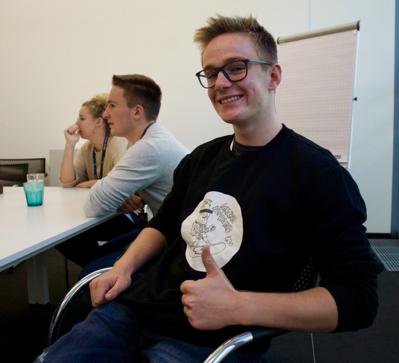Ein etwa 17-jähriger Schüler sitzt auf einem Stuhl in einem Konferenzraum. Er trägt ein schwarzes Shirt und hält den Daumen nach oben.