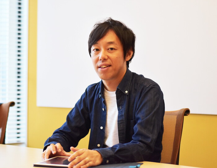 黄色い壁に白いボードを背景にミーティング用テーブルを前に座る男性。紺色のシャツ。