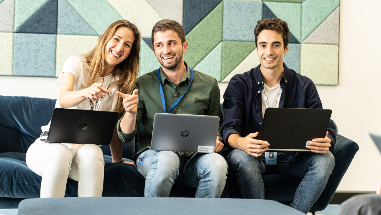 Silvia Cubero, Enrique Rodríguez y Pedro Mas preparando Prime Day.  Los tres están sentados en un sofá de color azul con los ordenadores sobre las piernas. Silvia va con pantalón y camiseta de color blanco. Enrique va con tejanos y una camisa verde camuflaje y Pedro con tejanos, una camiseta blanca y una camisa azul. Silvia y Enrique están hablando y Pedro mira a cámara. 