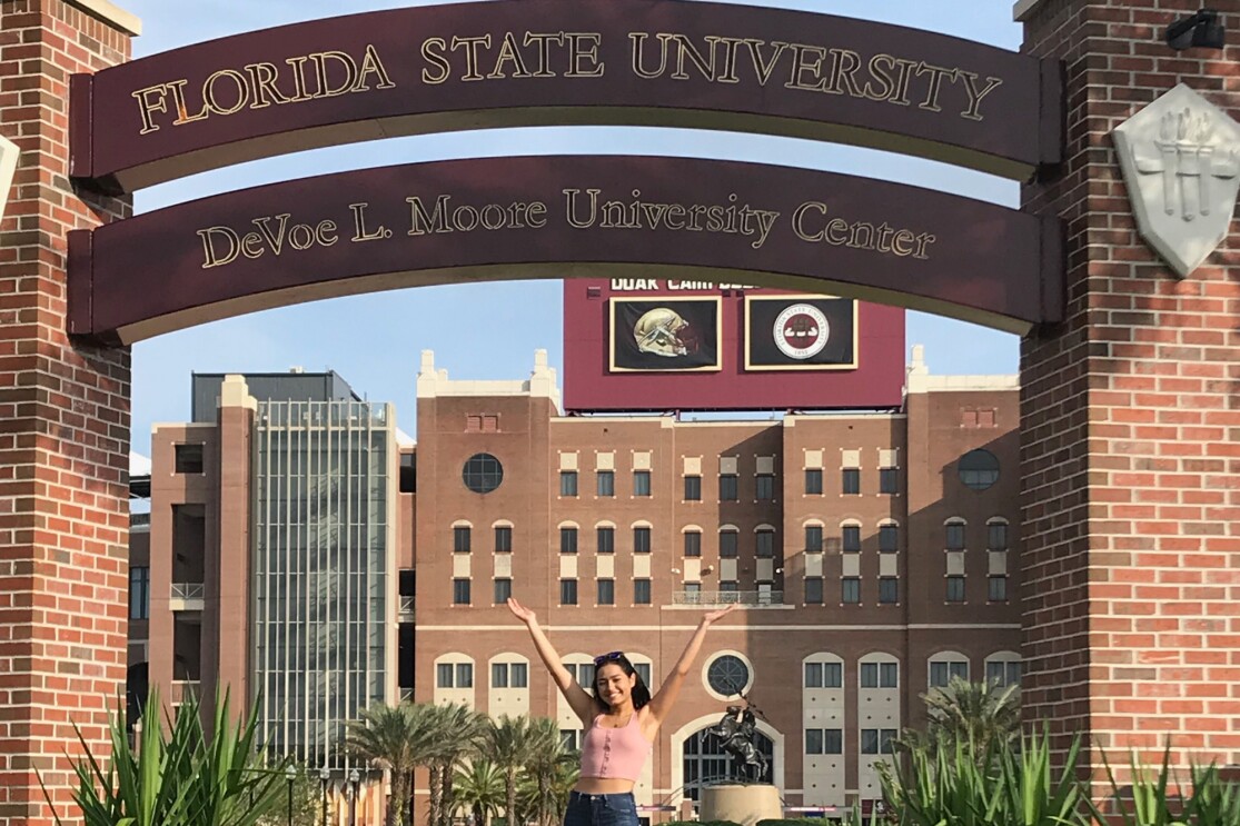 A young woman stands with her arms raised under a Florida State University sign.
