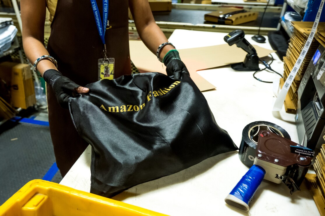 A person photographed from the torso down holds a black satin bag labeled with the words "Amazon Fashion." The image also contains packing tape and a scanner. Boxes move along a conveyor belt in the background.