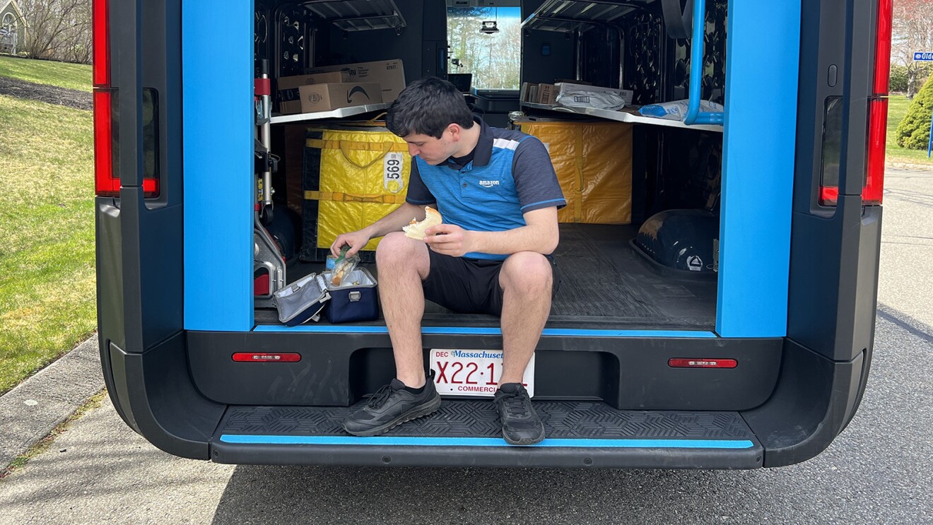 An Amazon delivery driver sits on the back of a Rivian van and eats lunch.