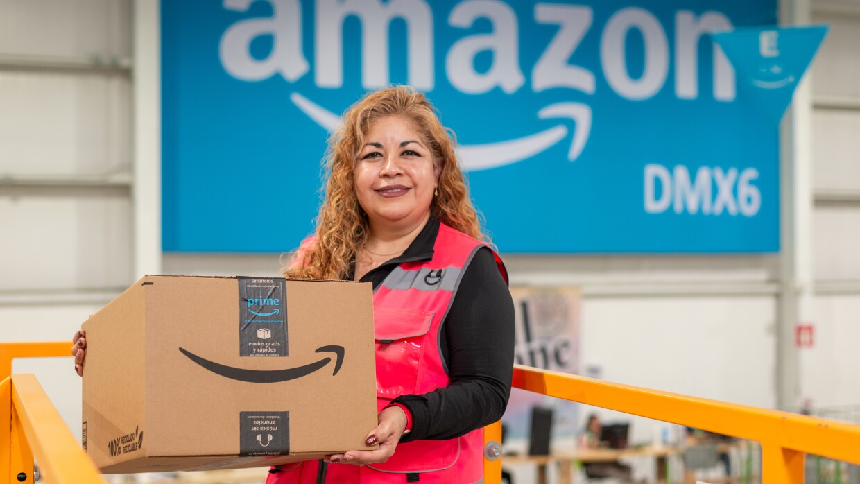 Trabajadora de Amazon cargando una caja delante de un poster que pone Amazon DMX6.