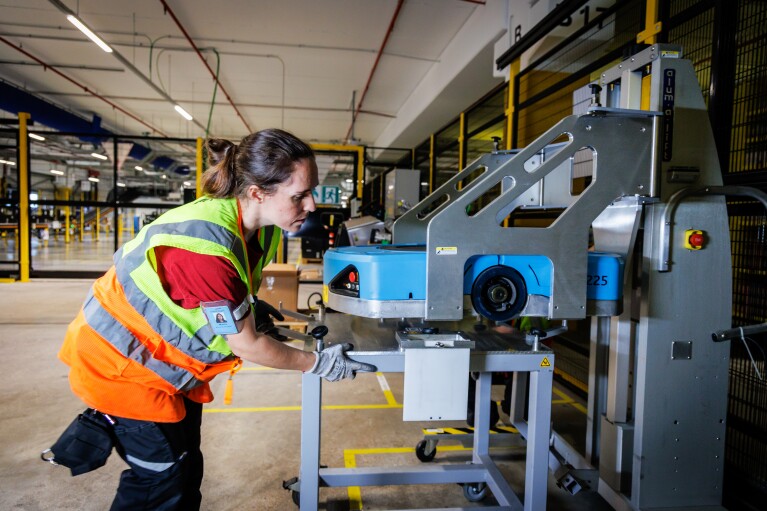 Dentro de un centro logístico, está una mujer con un elevador para reparar un robot de color azul. Está de pie, lleva el pelo recogido con un moñparrodillada mirando un robot está colocado en una mesa. Ella tiene el pelo oscuro largo y recogido en una cola. Van con un camiseta negra de manga corta y un chaleco amarillo. 