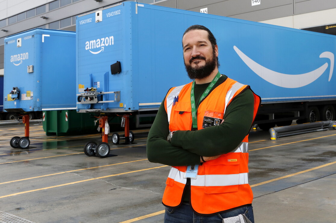 Sergio Amador con los brazos cruzados, un chaleco naranja, mira a cámara. Tiene barba y el pelo largo negro, recogido con una cola. De fondo están dos remolques azules con la sonrisa de Amazon. 