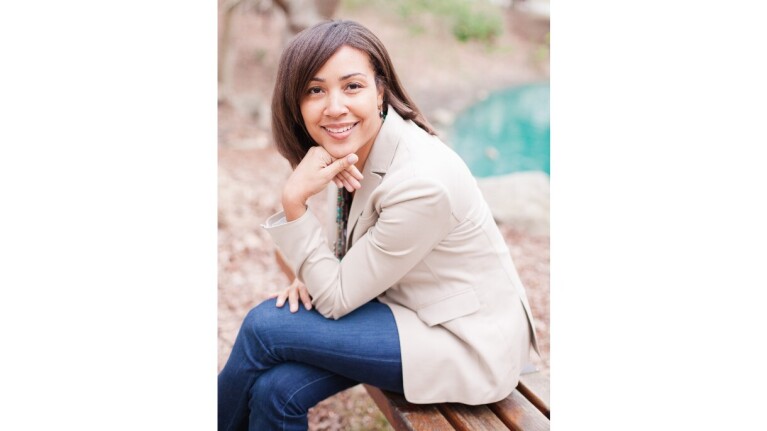 Melanie LeGrande sits on a bench and poses outside wearing jeans a tan blazer.